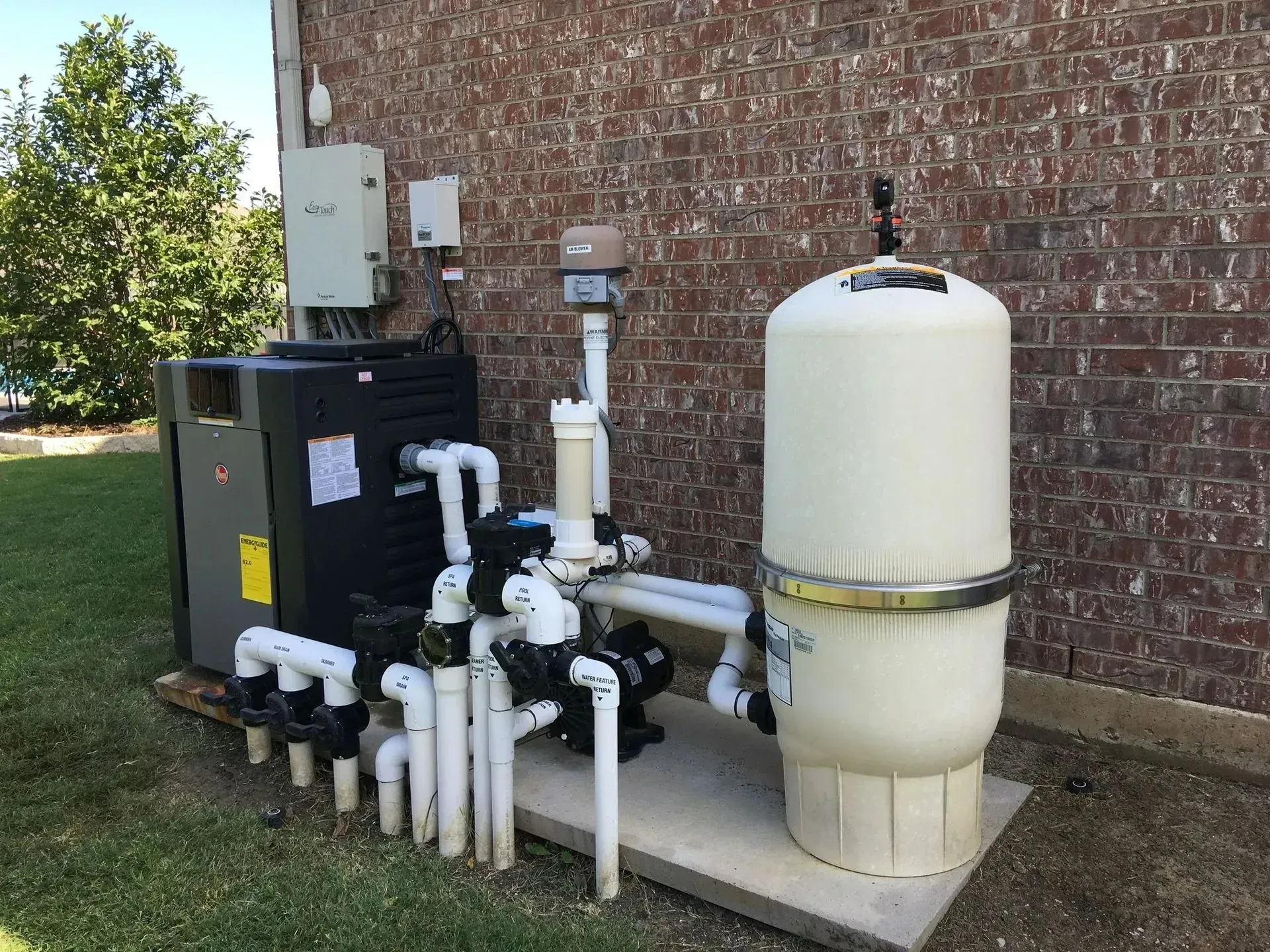 Pool equipment including heater, filter, and plumbing against a brick wall and grass.