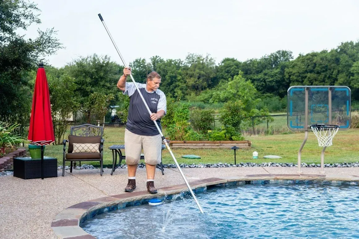 Man cleaning a blue swimming pool with a long-handled pole. Outdoor setting with trees and a basketball hoop.