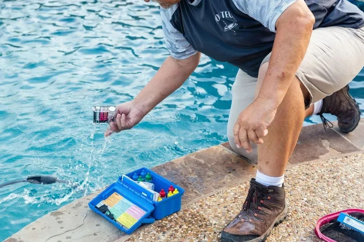 Person kneels by water, dropping a small device into the blue water. A blue box with toys sits next to them.