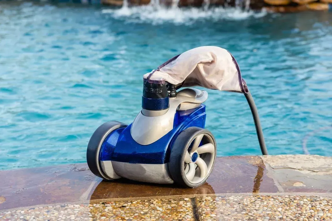 Blue and white robotic pool cleaner on the edge of a pool with a white waterfall in the background.