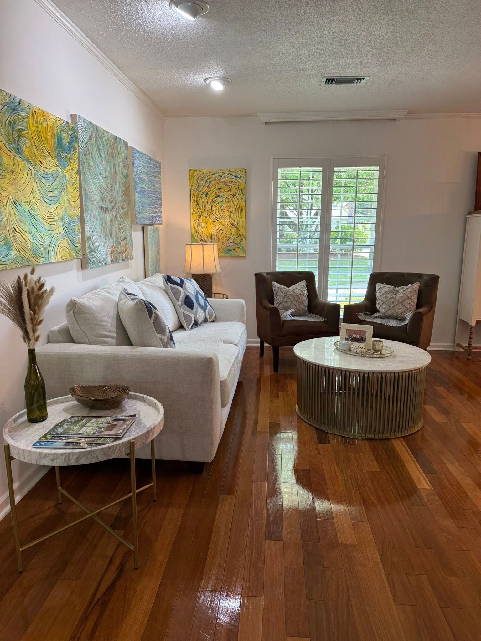 Living room with white sofa, brown armchairs, colorful art, and a marble coffee table.