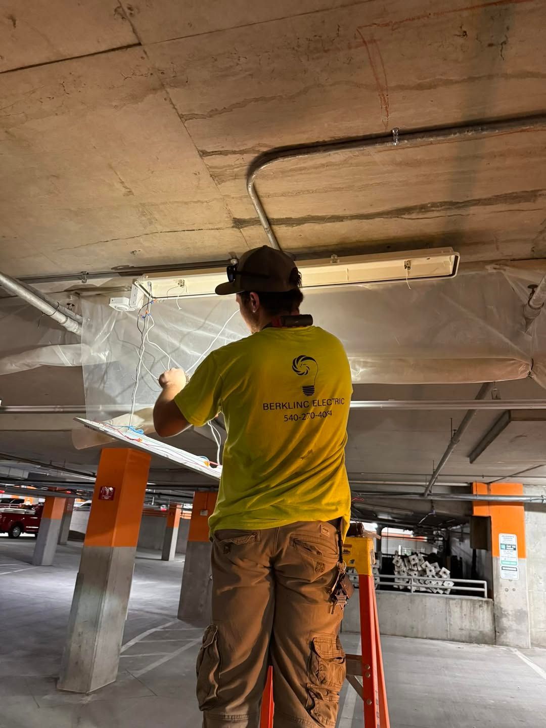 Man in yellow shirt on a ladder, working on ceiling in a parking garage.