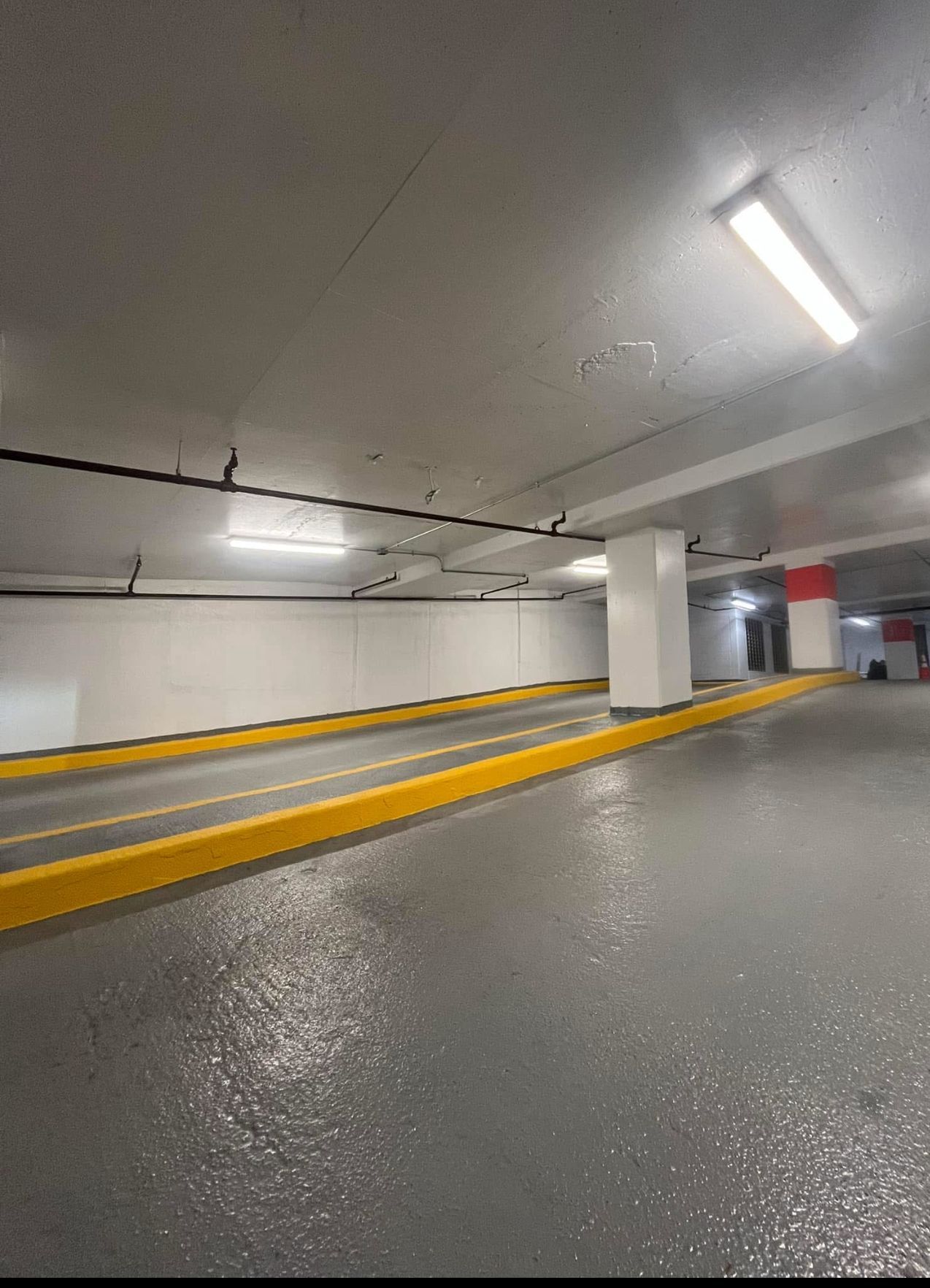 Empty parking garage with wet, gray floor and yellow curbs. Overhead lights illuminate white walls and a white pillar.