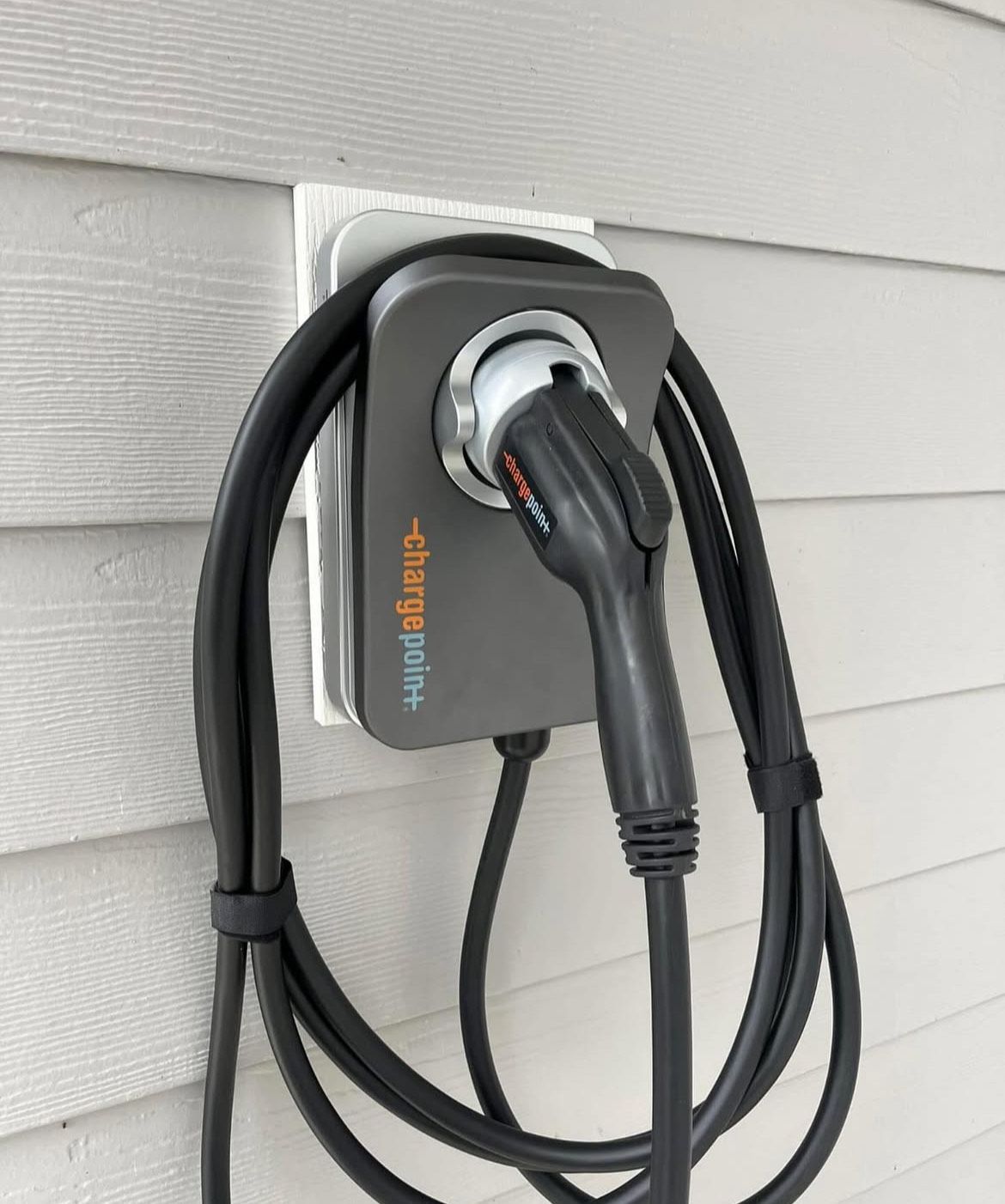 Electric vehicle charger mounted on a house exterior. Gray charger with black cable coiled beside it.