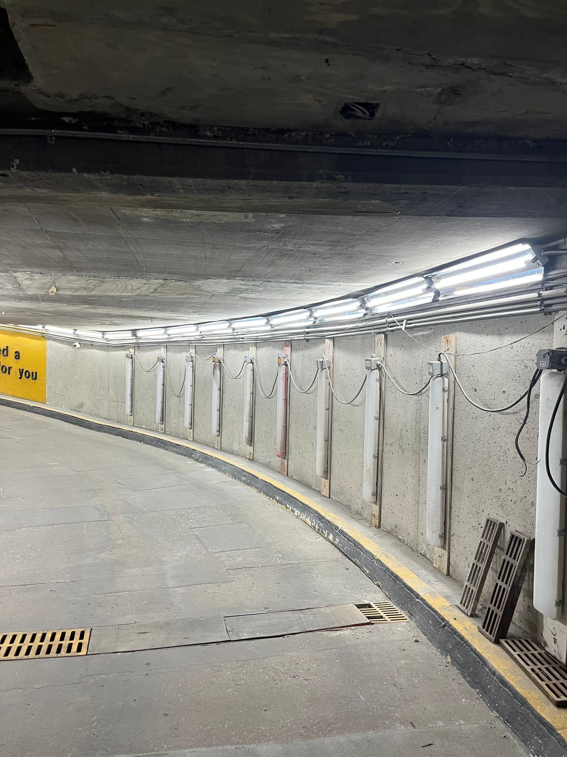 Curved, dimly lit parking garage entrance with concrete walls, overhead fluorescent lights, and a yellow sign.