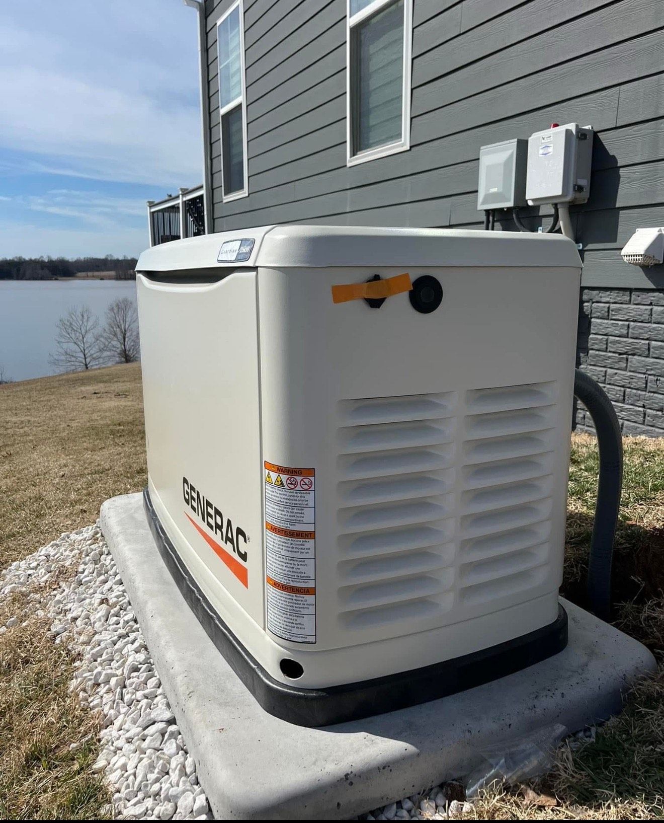 Generator unit on a concrete pad next to a house by a body of water on a sunny day.