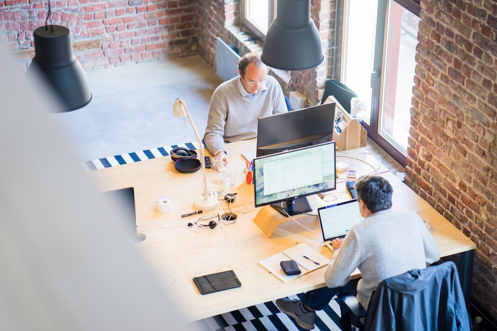 Drie mensen werken aan bureaus in een licht kantoor met bakstenen muren, met computers, laptops en papieren.