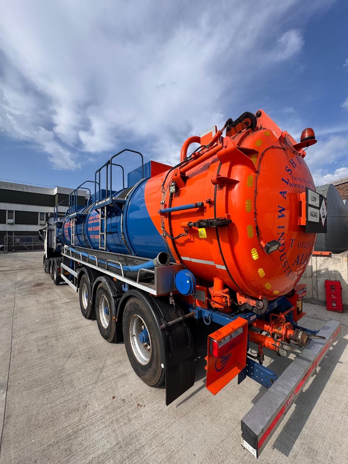 Orange and blue industrial tank truck parked on a concrete surface against a light blue sky.