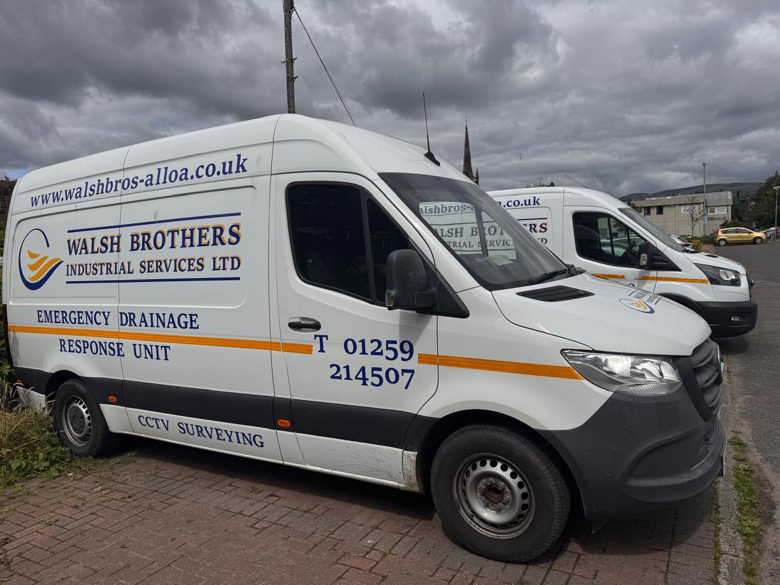Two white Walsh Brothers industrial service vans parked on a road with company details and contact information.