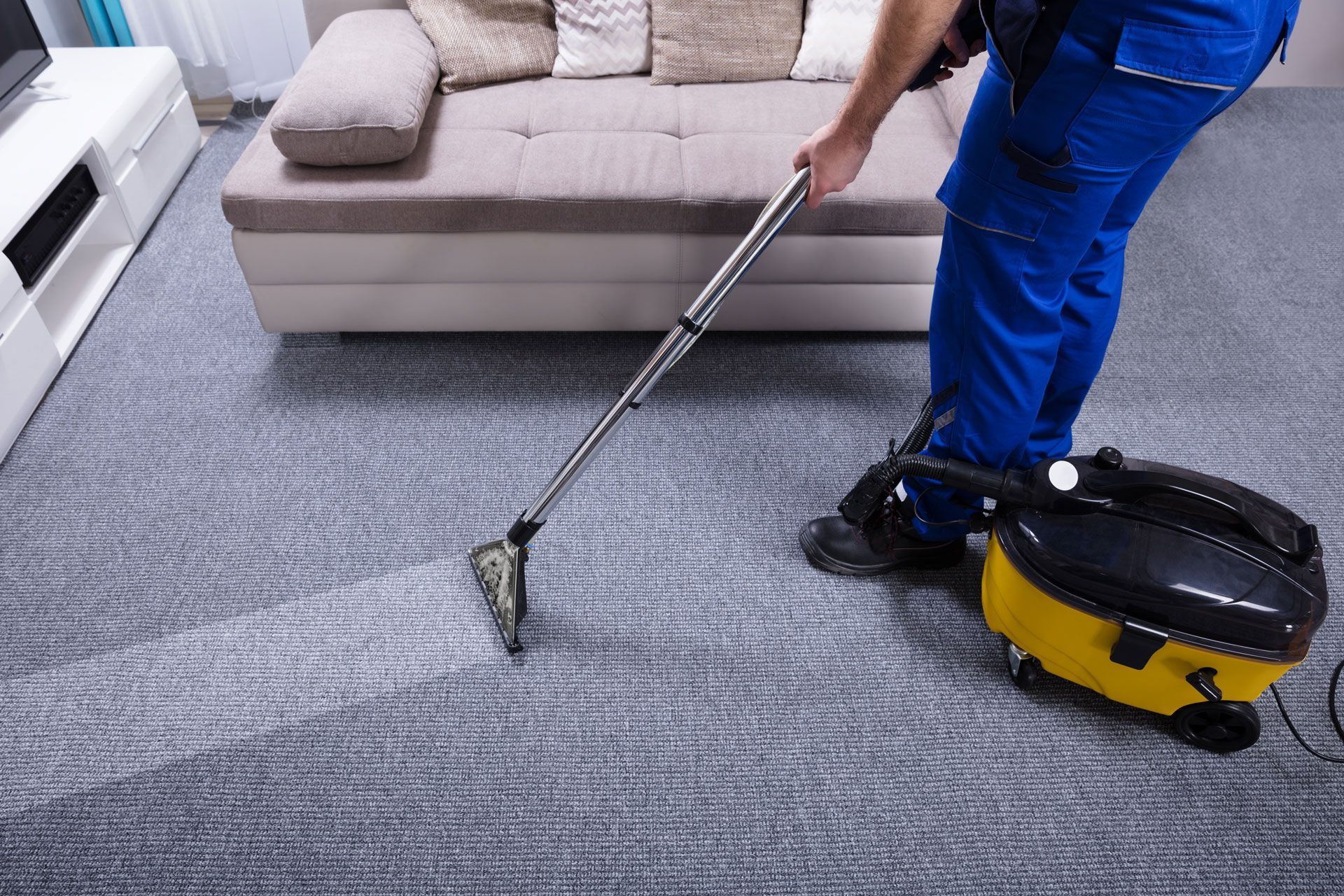 Person in blue cleaning carpet with a yellow vacuum cleaner in a living room.