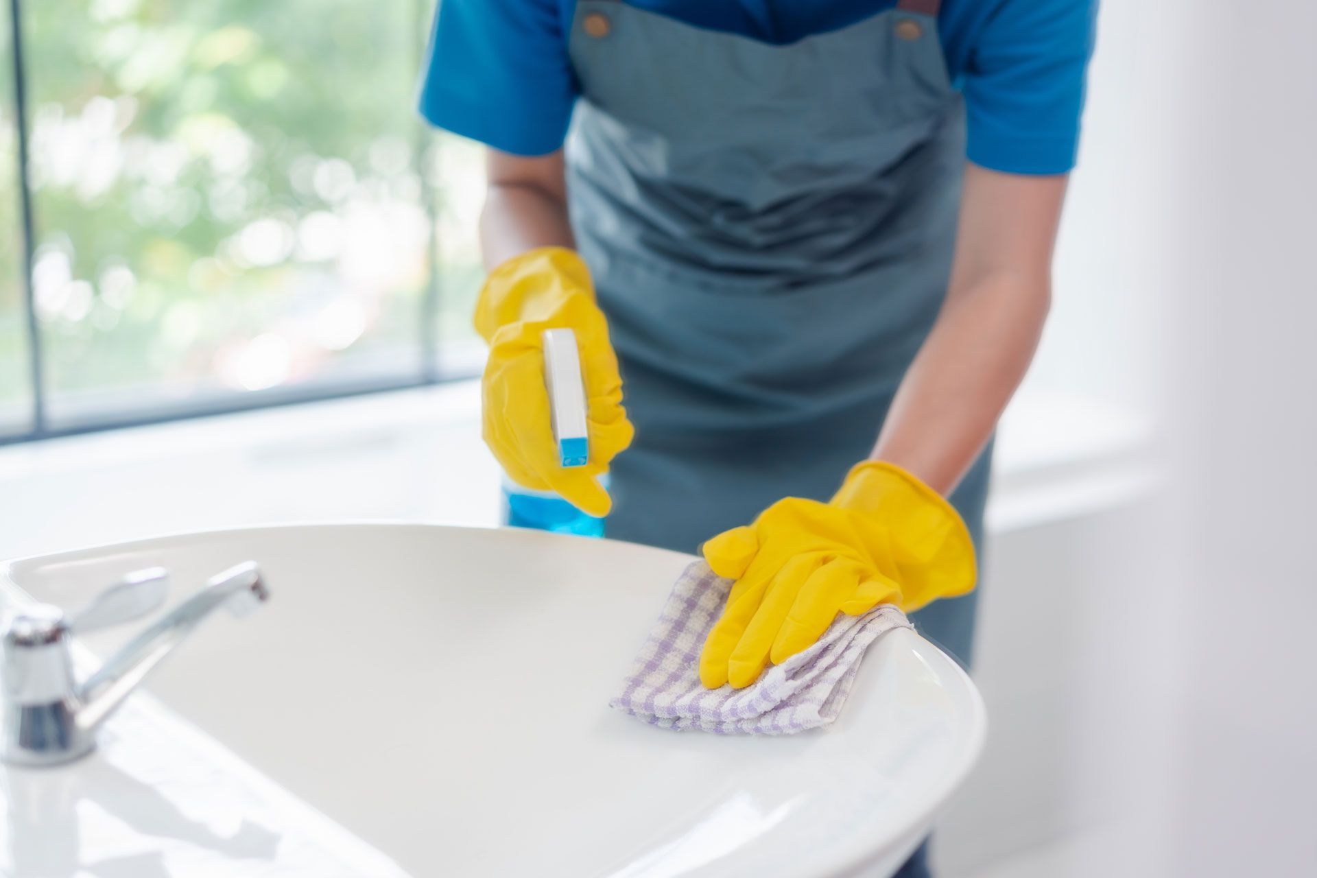 Person wearing gloves and apron cleaning a white sink in a bathroom with a window in the background.