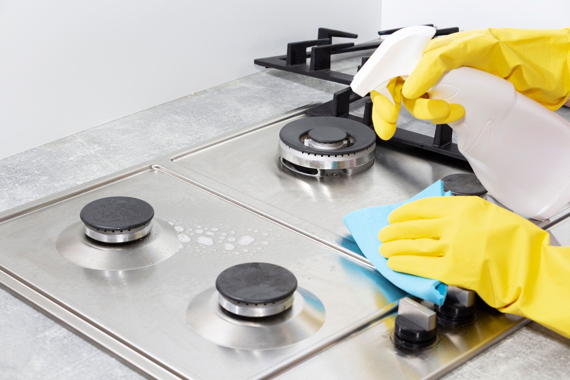 Hands in yellow gloves cleaning a stainless steel gas stovetop with spray and a blue cloth.