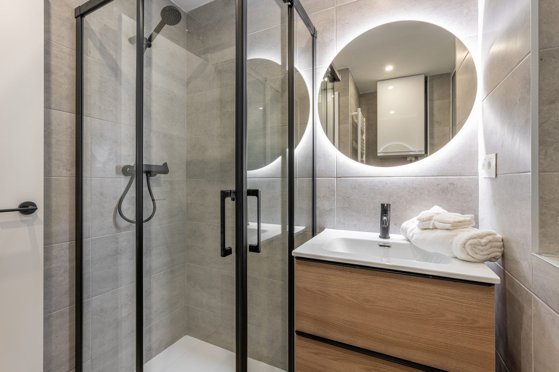 Modern bathroom with a glass shower, wooden vanity, and a round mirror with lights.