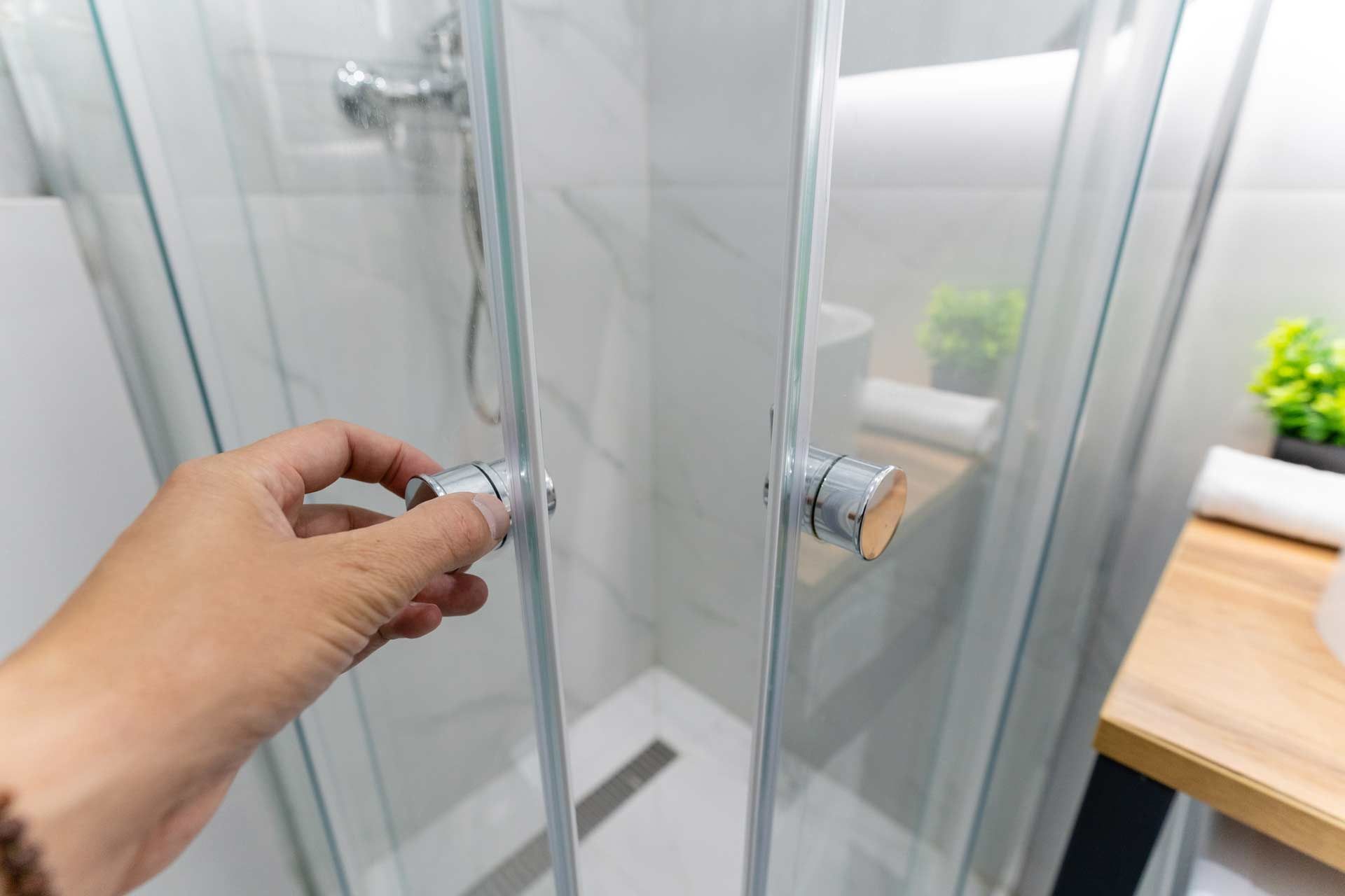 Hand opening a glass shower door with chrome handles. Bathroom interior.