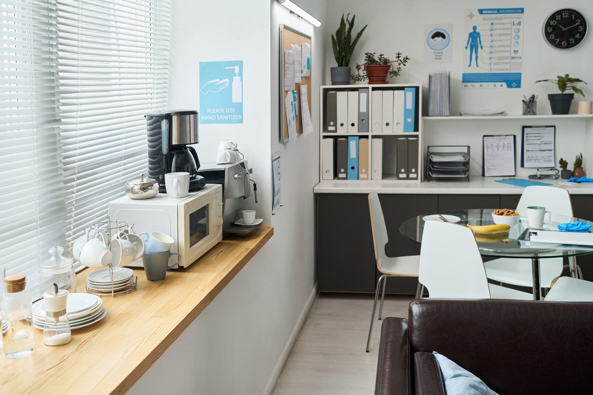 Office breakroom: microwave, coffee maker, window, small table with chairs, and a brown sofa.