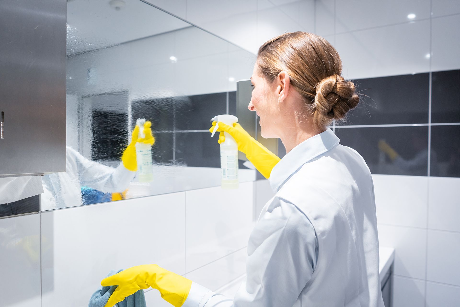 Woman in yellow gloves sprays and wipes a bathroom mirror.