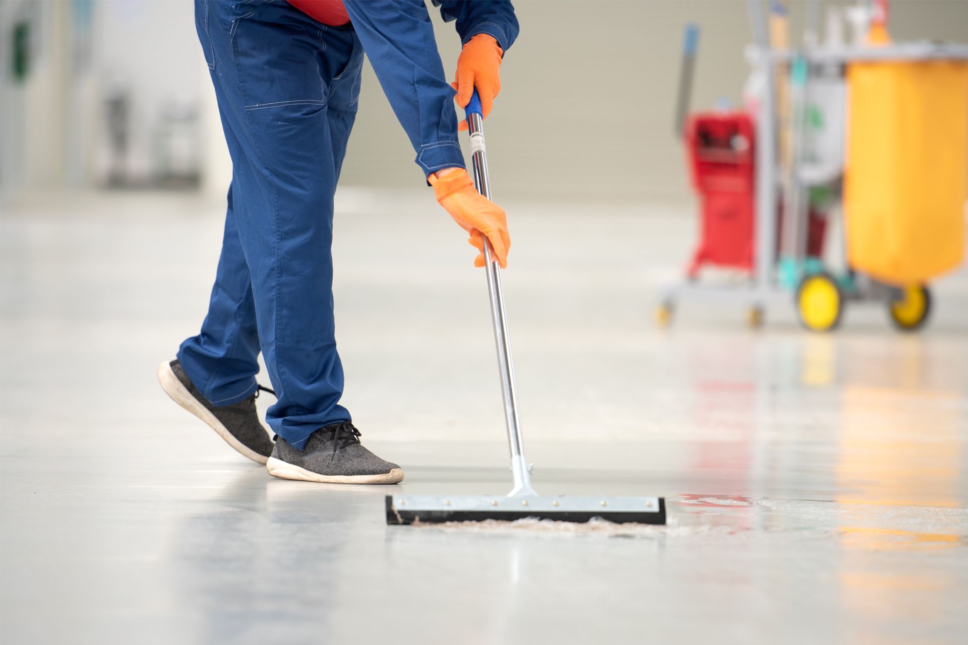 Person in blue coveralls mops a wet floor. Orange gloves. Cleaning cart in background.
