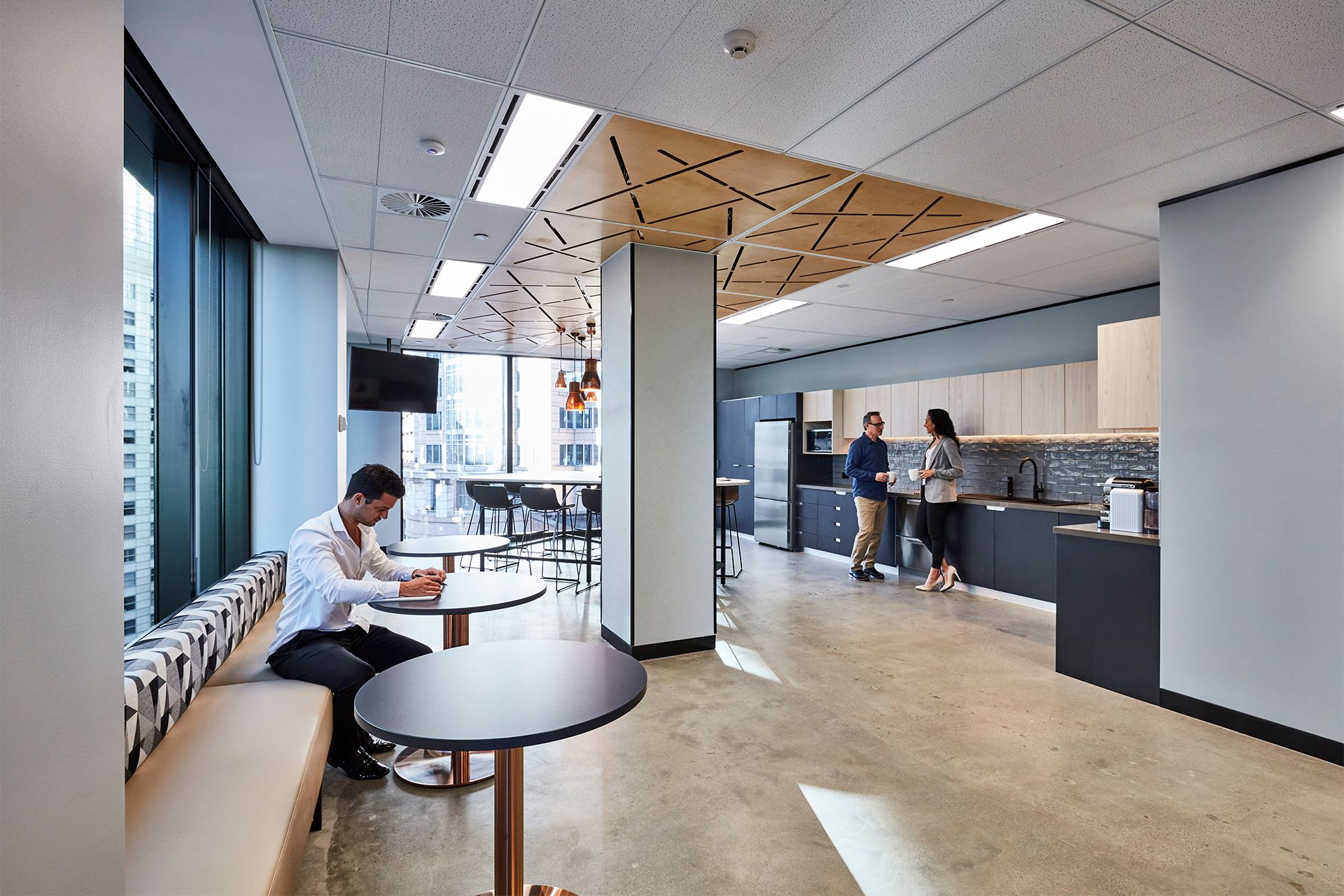 Office interior with people in a seating area and kitchen; one person writing, others standing near a counter.