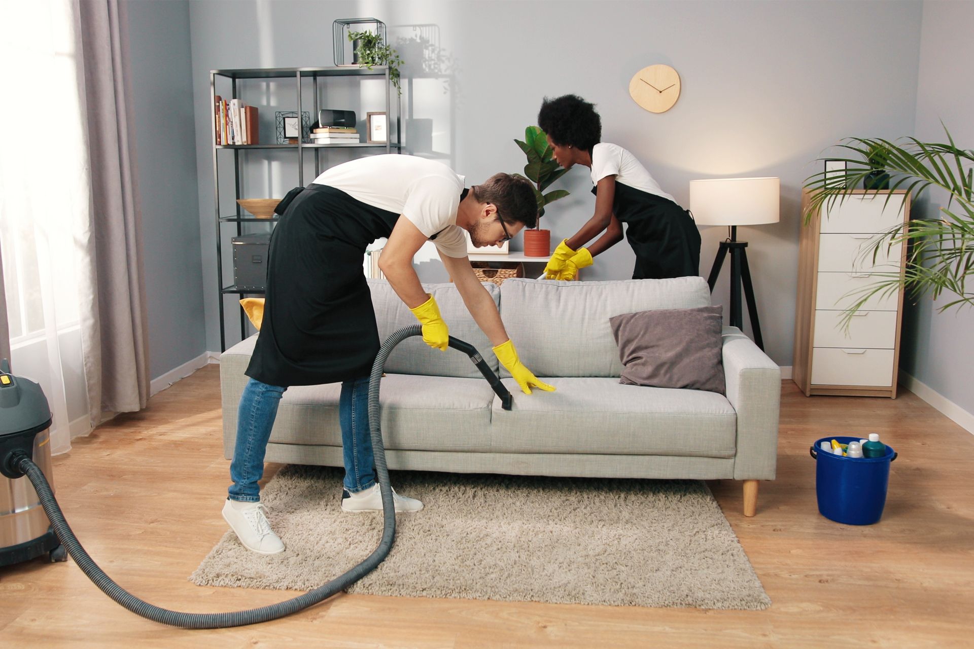 Two people wearing aprons and gloves vacuum a sofa in a living room.
