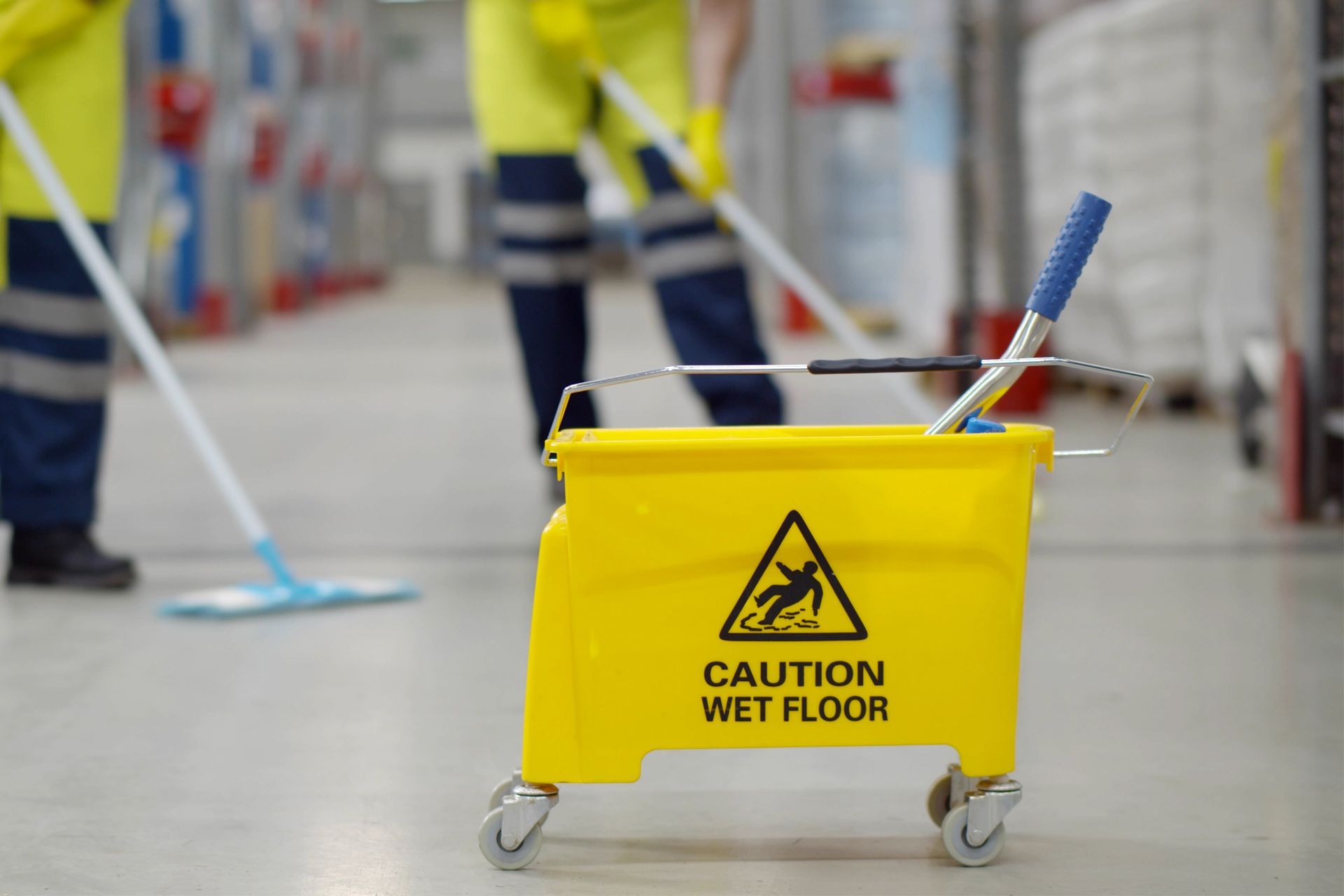 Yellow wet floor sign with mop bucket, workers mopping floor in a warehouse.