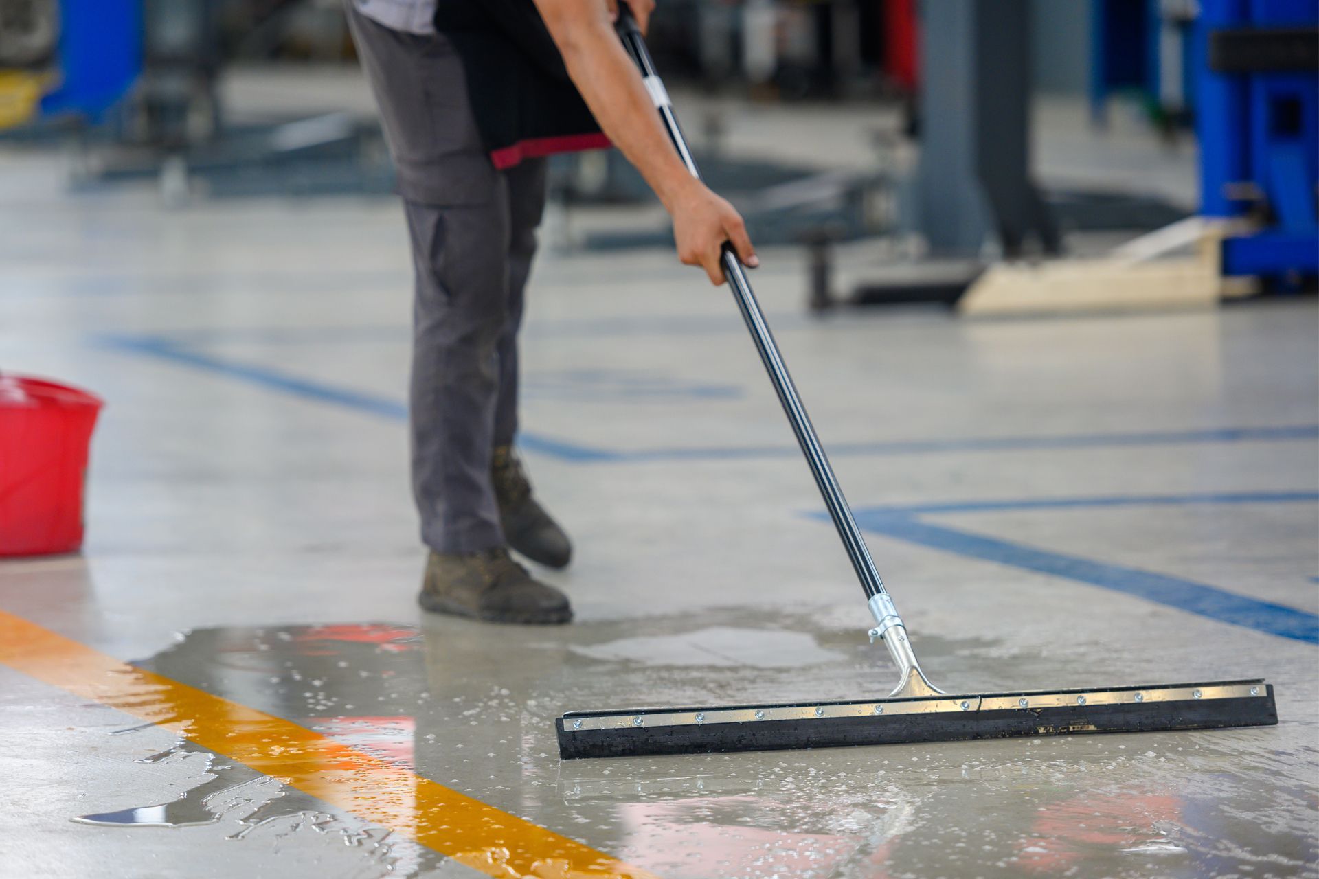 Person using a squeegee to clean a wet floor in a workshop, next to a red bucket and yellow line.