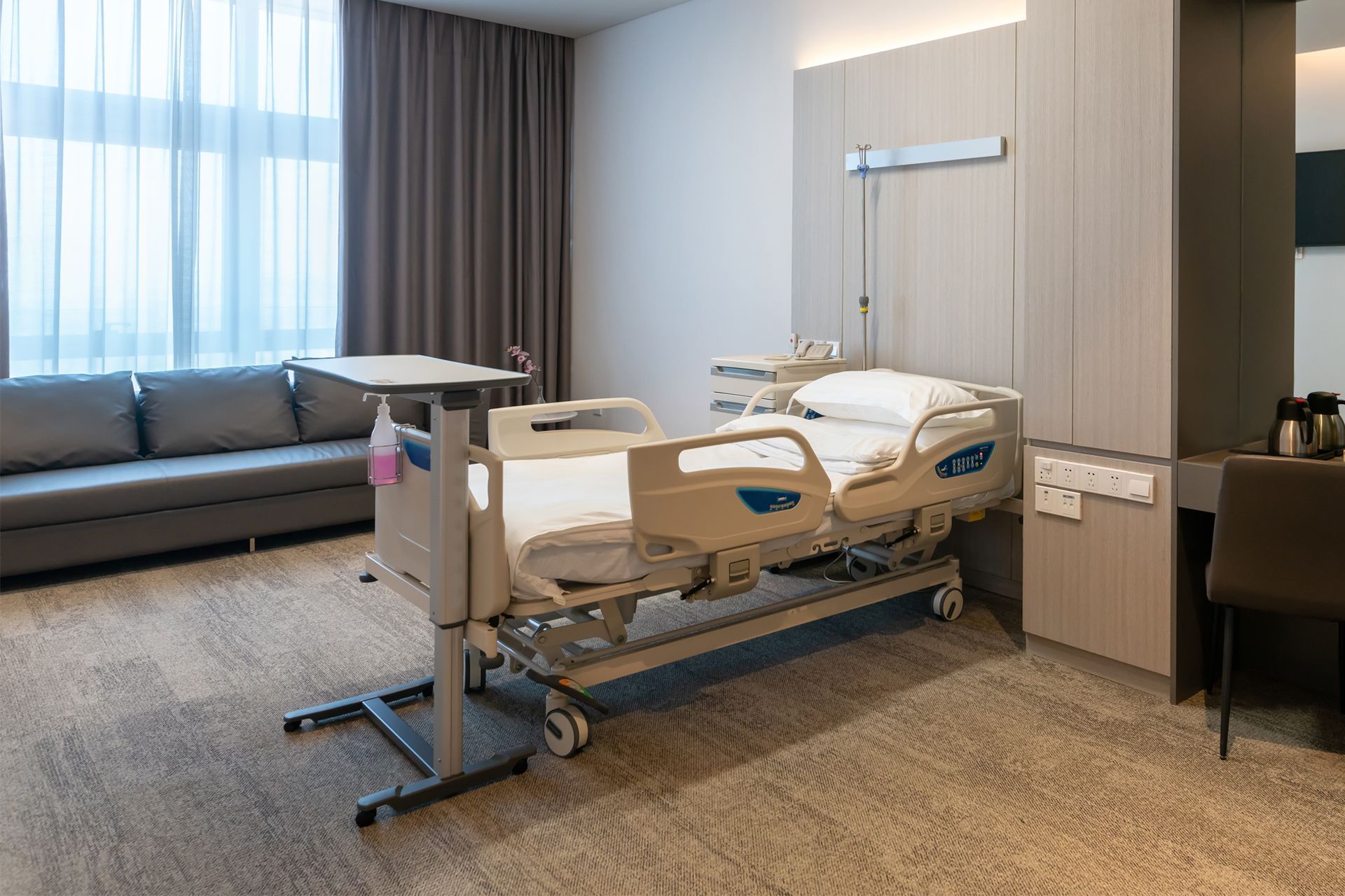 Hospital room with adjustable bed, sofa, and desk. Soft lighting and neutral tones.