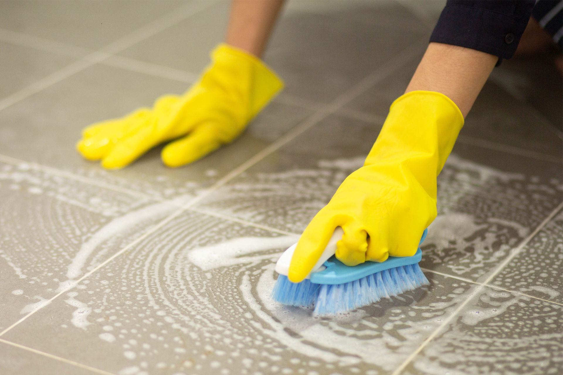 Hands in yellow gloves scrubbing a tiled floor with a blue brush, surrounded by soapy suds.
