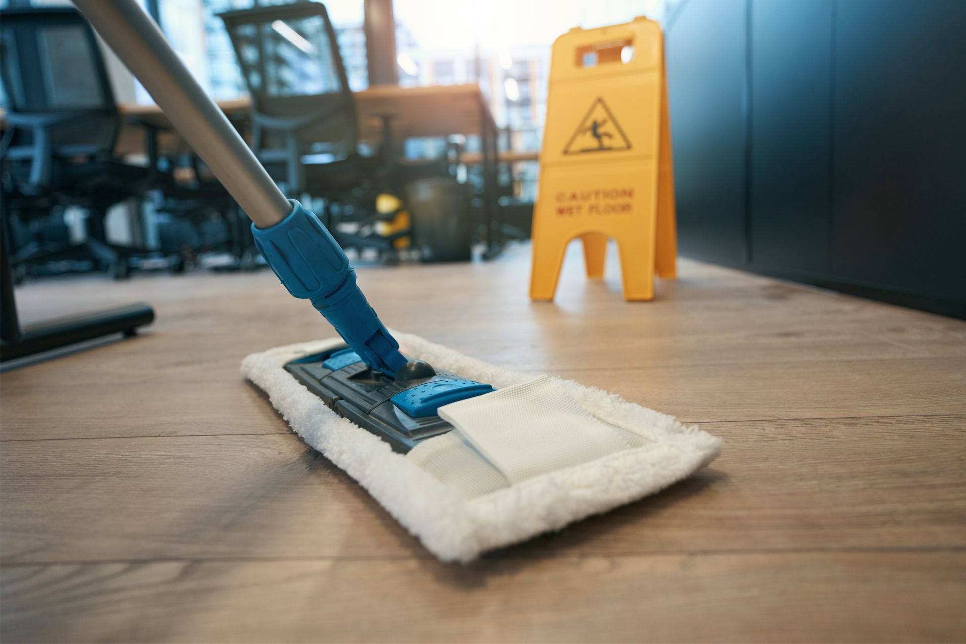A mop cleaning a wooden floor in an office, with a wet floor sign in the background.