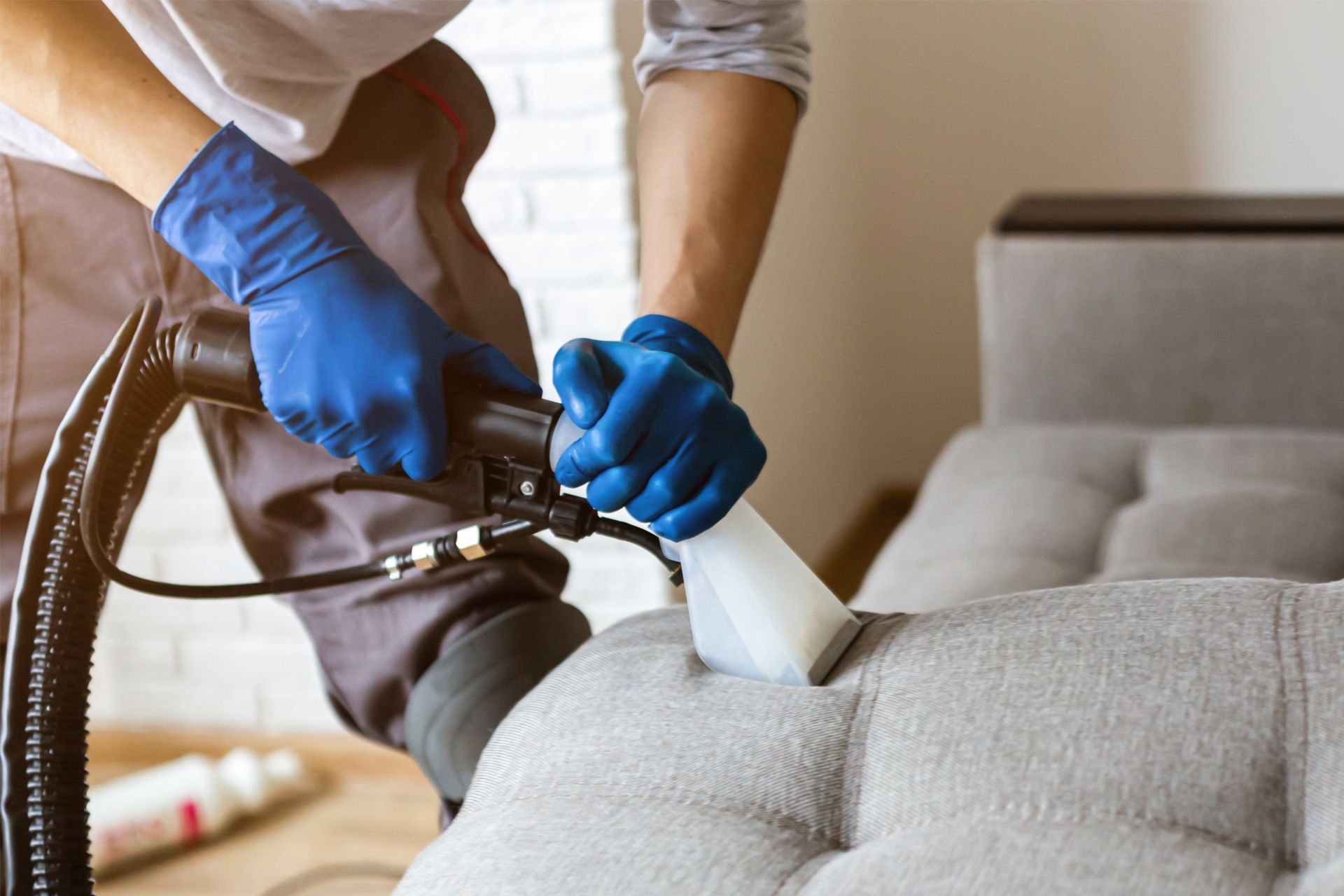 Person in blue gloves cleaning a gray sofa with a steam cleaner.