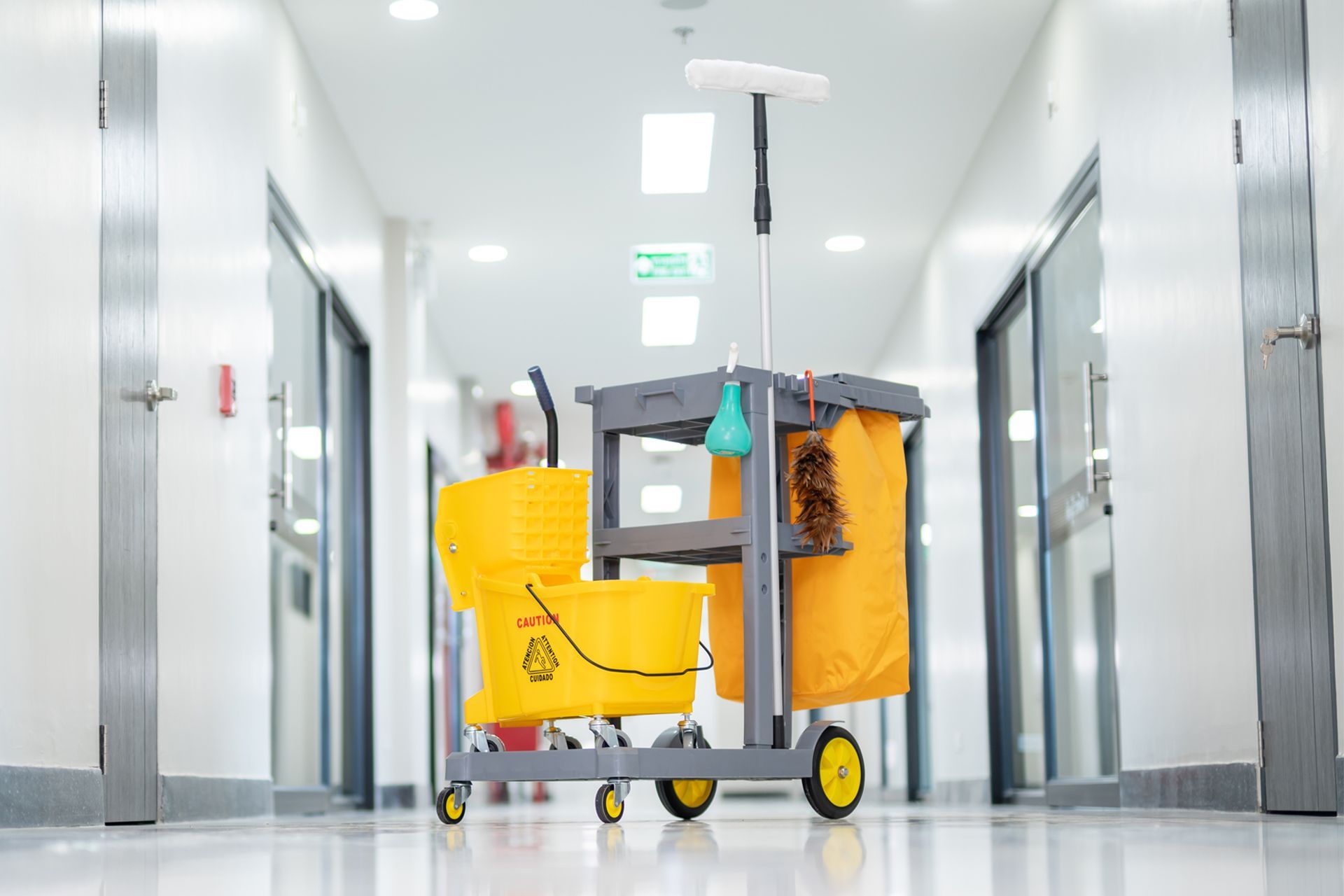 Cleaning cart in a brightly lit hallway; yellow buckets, mop, and storage bag.