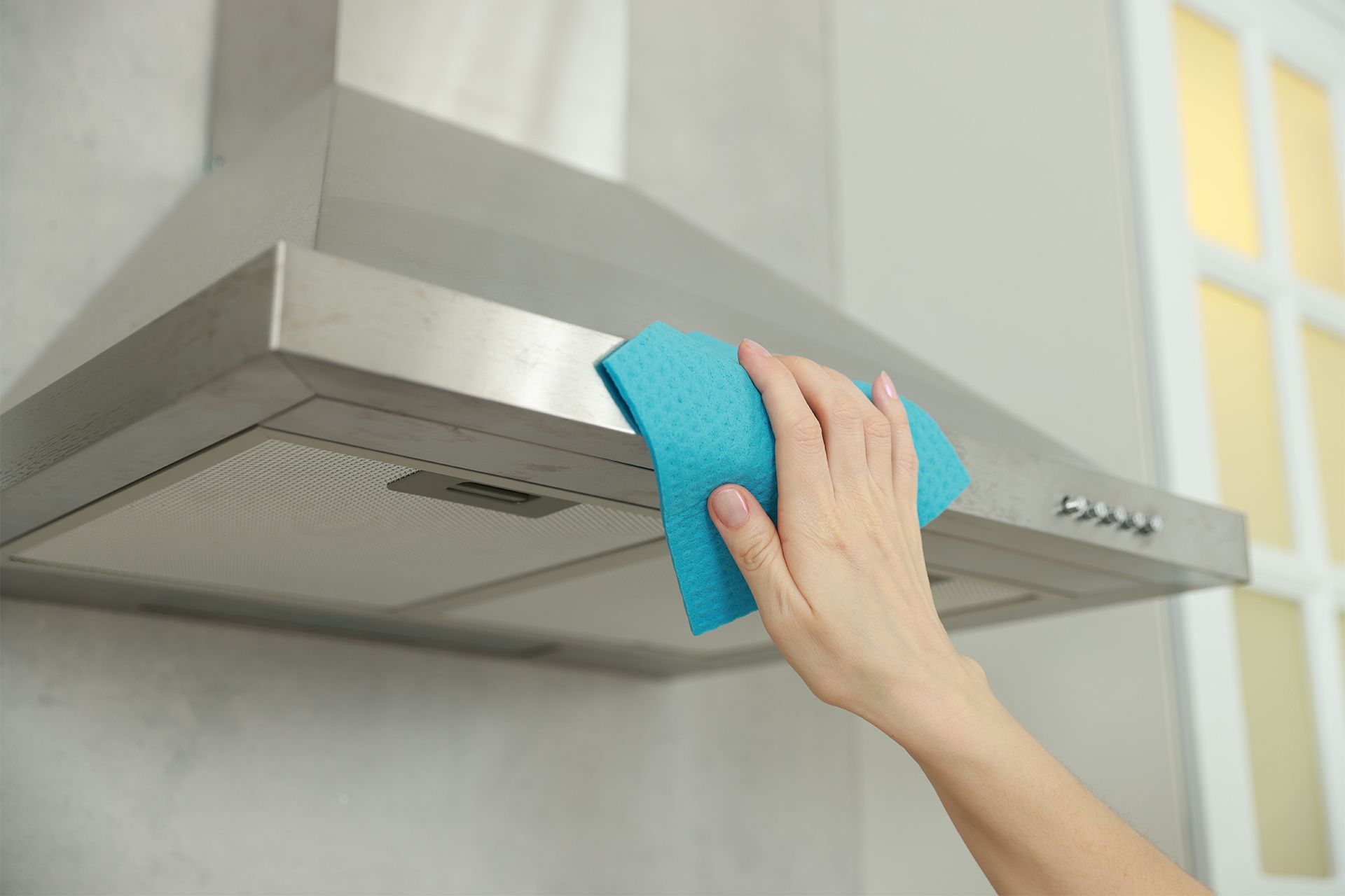 Hand wiping down a stainless steel range hood with a blue cloth in a kitchen.