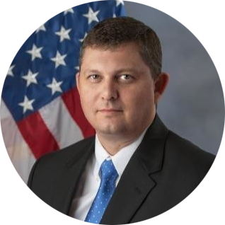 Portrait of a man in a dark suit and blue tie before a U.S. flag backdrop
