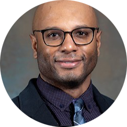 Portrait of a man wearing glasses, a dark suit, and patterned tie against a gray background