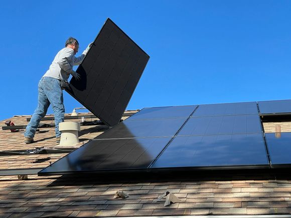 A worker installs black solar panels onto the shingled roof of a house under a clear blue sky.