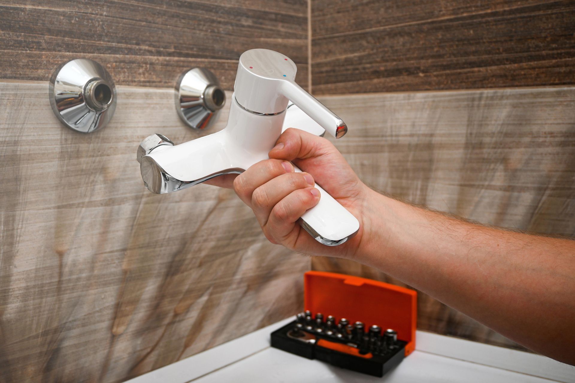 A hand holds a white and chrome shower faucet in front of a tiled wall with two exposed pipe fittings and a tool kit.
