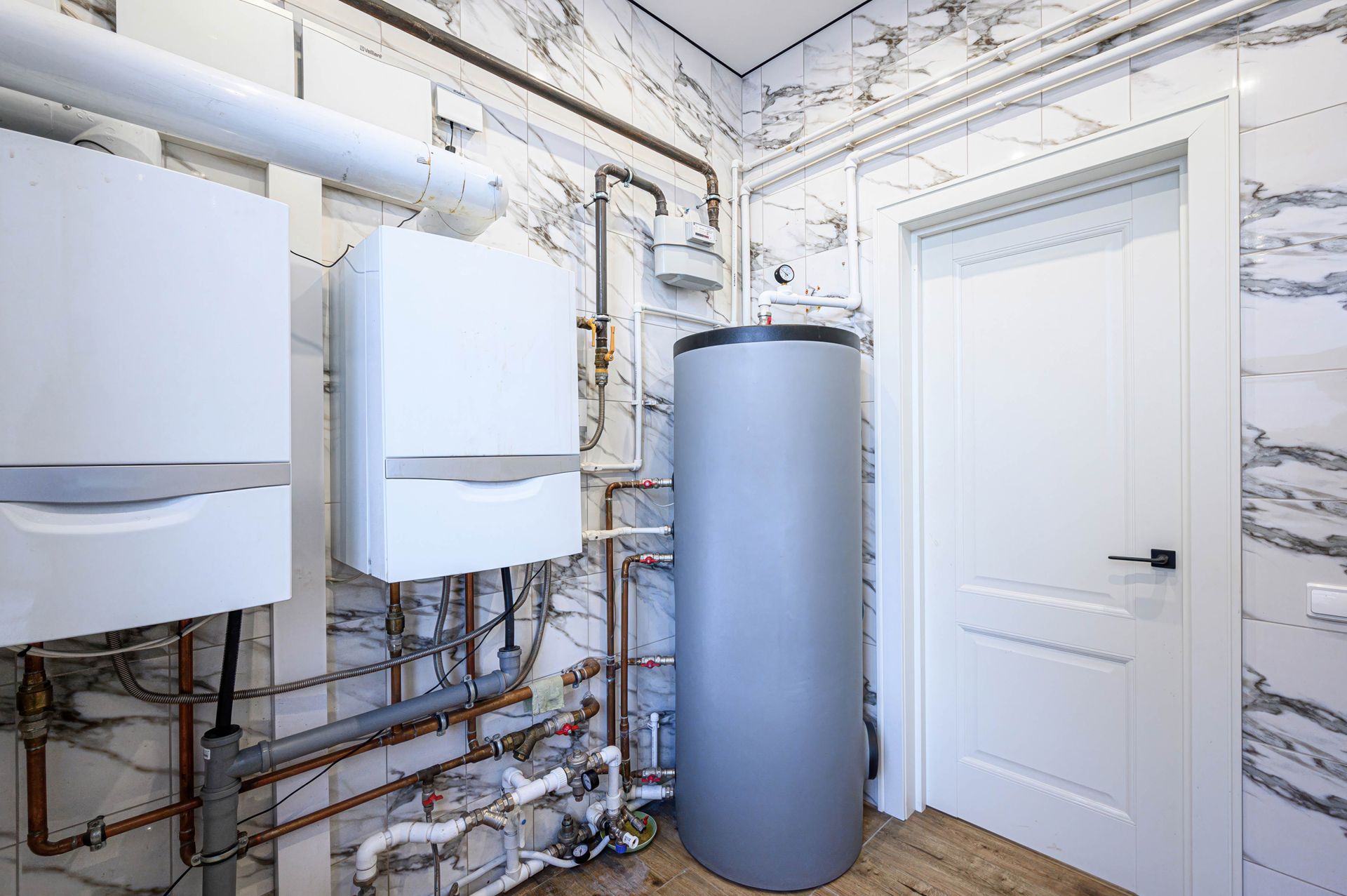 Two white wall-mounted boilers and a large grey water tank in a utility room with marble-patterned walls and a white door.