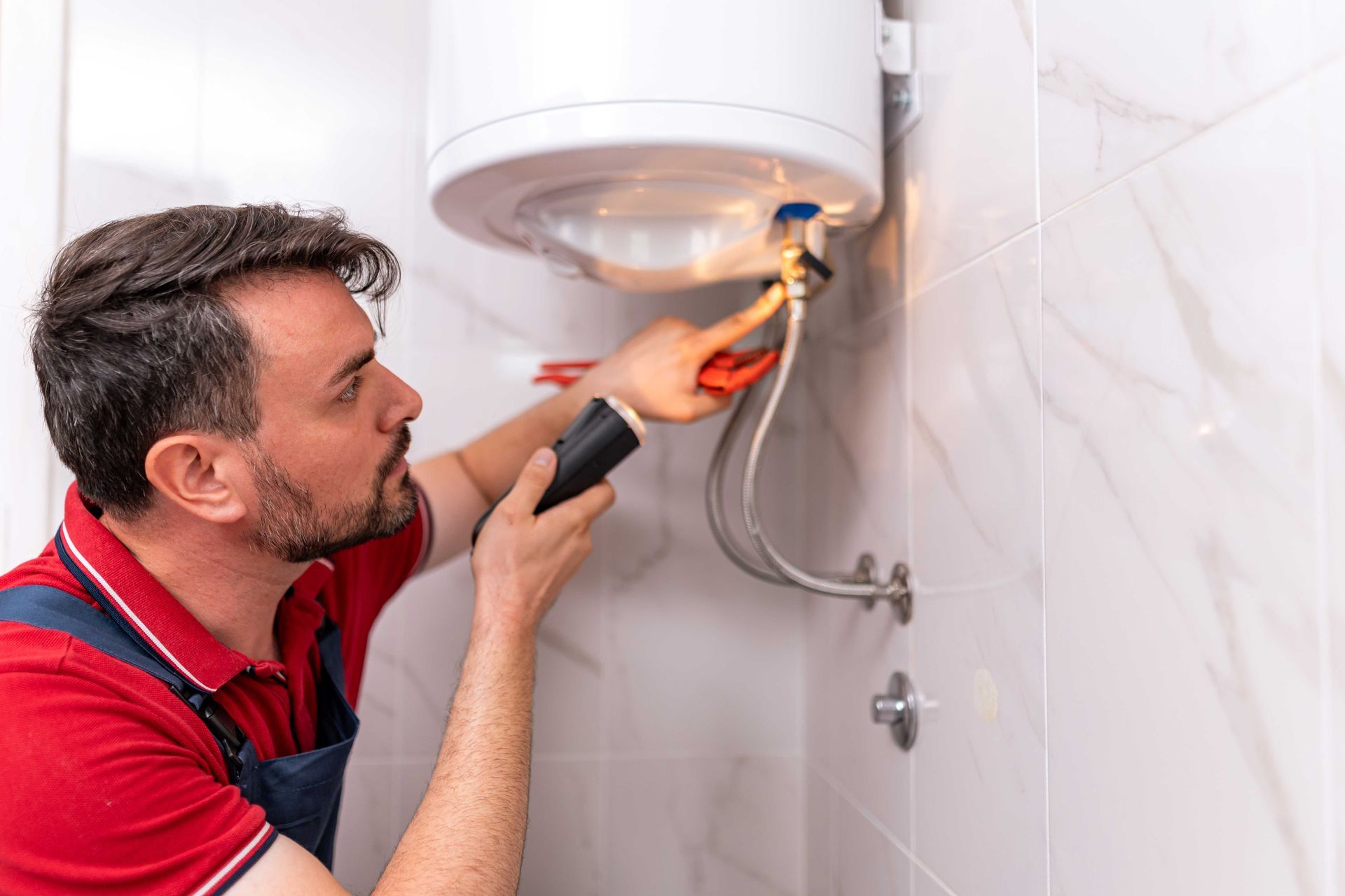 A person in a red shirt and work overalls inspects a wall-mounted water heater with a flashlight.
