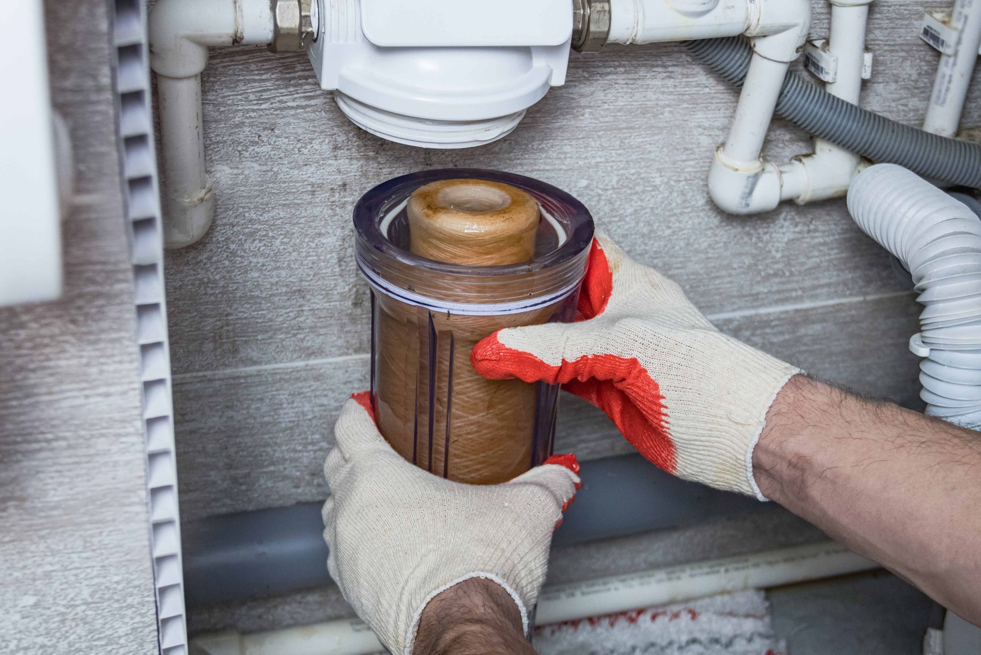 Hands wearing work gloves installing a replacement water filter cartridge into its housing under a sink.
