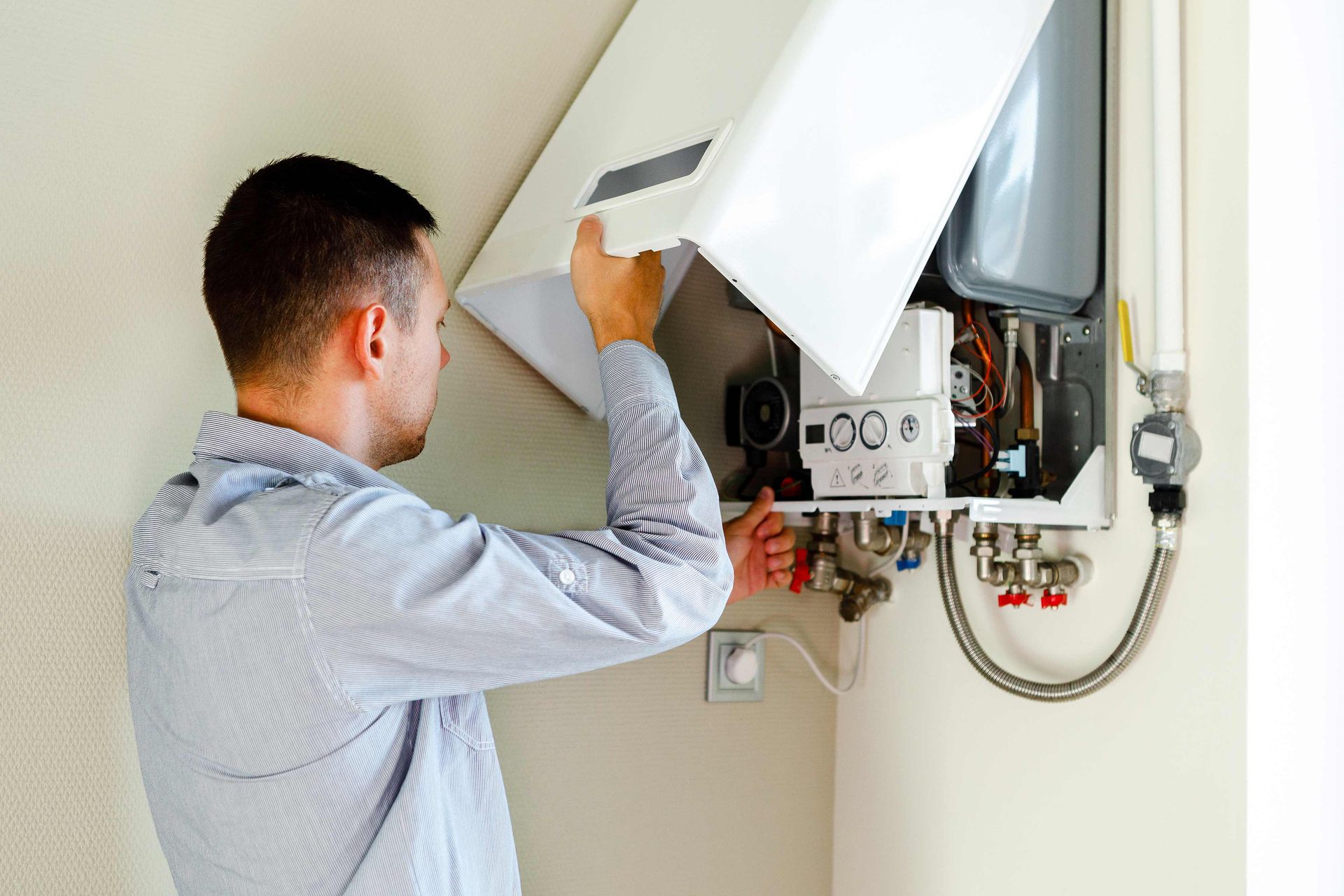 A technician in a light-colored shirt works on an open wall-mounted boiler unit indoors.