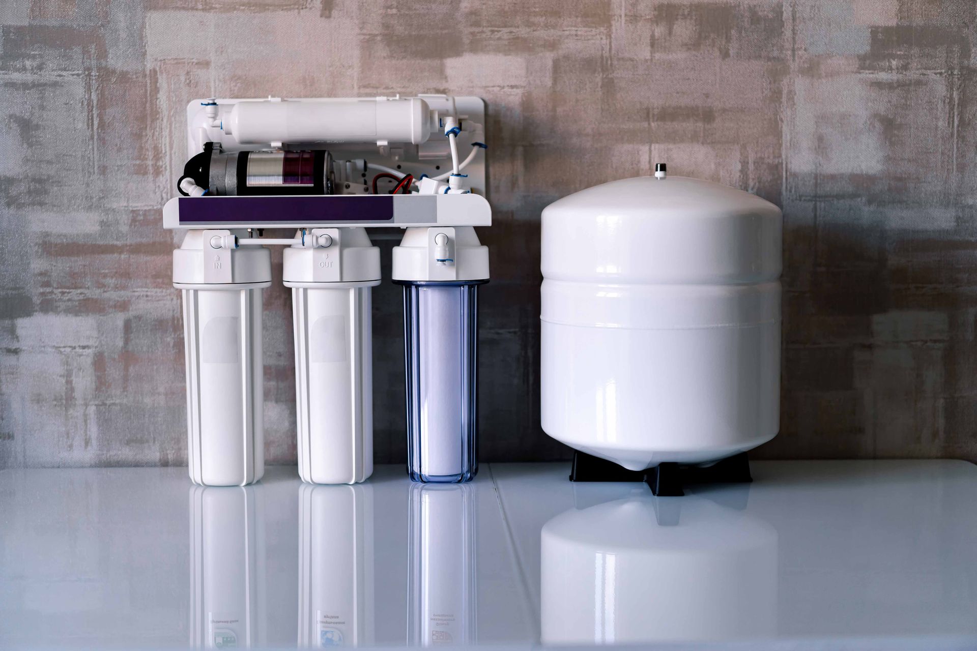 A white water filtration system with three filter housings and a large storage tank sits against a textured wall.