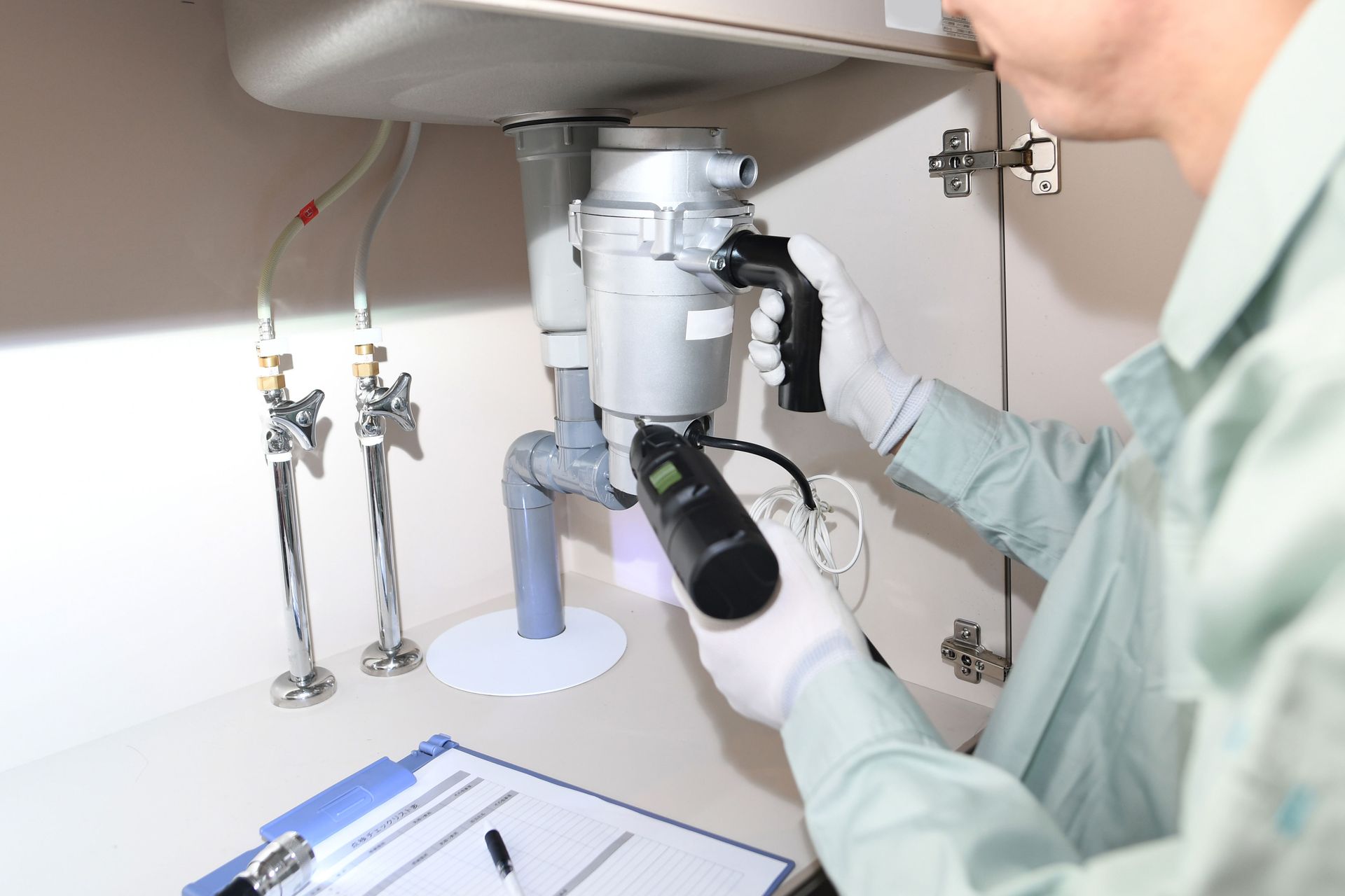 A technician in a light-colored uniform inspecting plumbing under a sink using a handheld diagnostic tool.