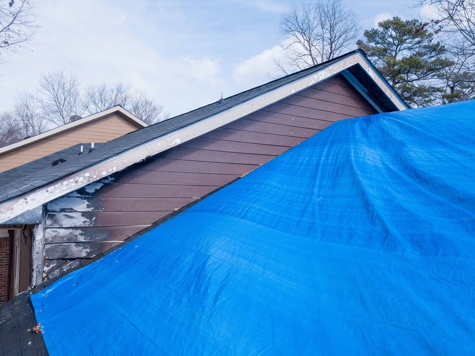 A blue tarp covering part of a residential roof where brown shingles are missing or damaged.