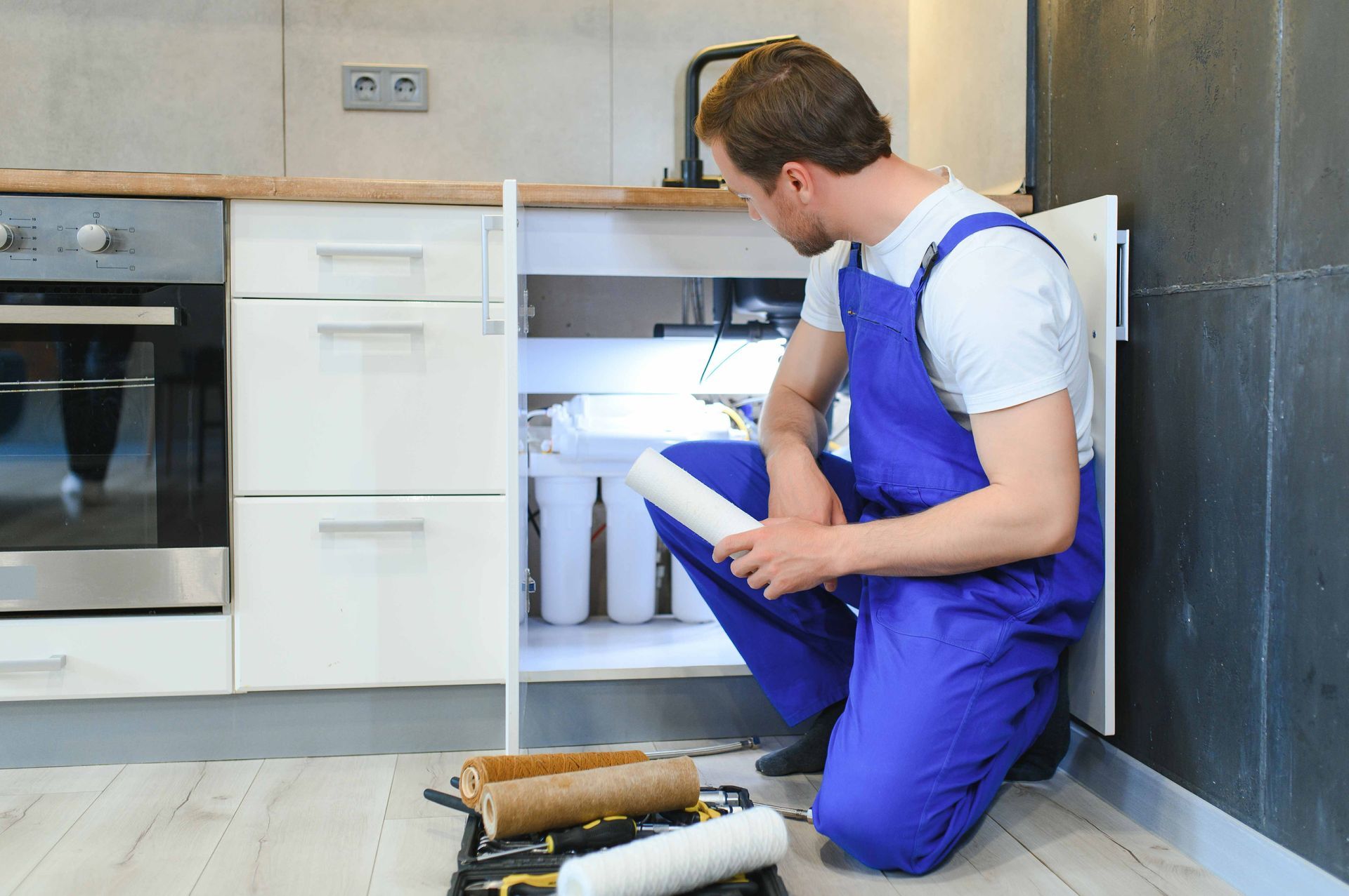 A technician in blue coveralls kneels under a kitchen counter, holding a replacement water filter near an installed unit.