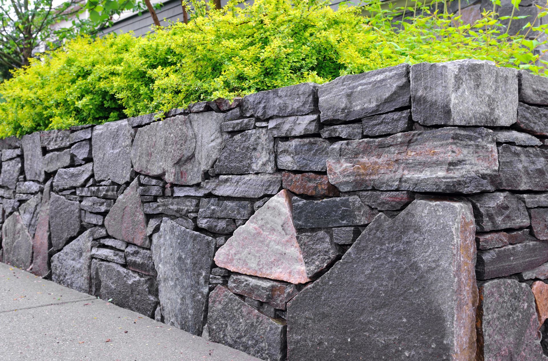 Natural stone retaining wall lining a sidewalk with layered rock blocks and green shrubs on top. 