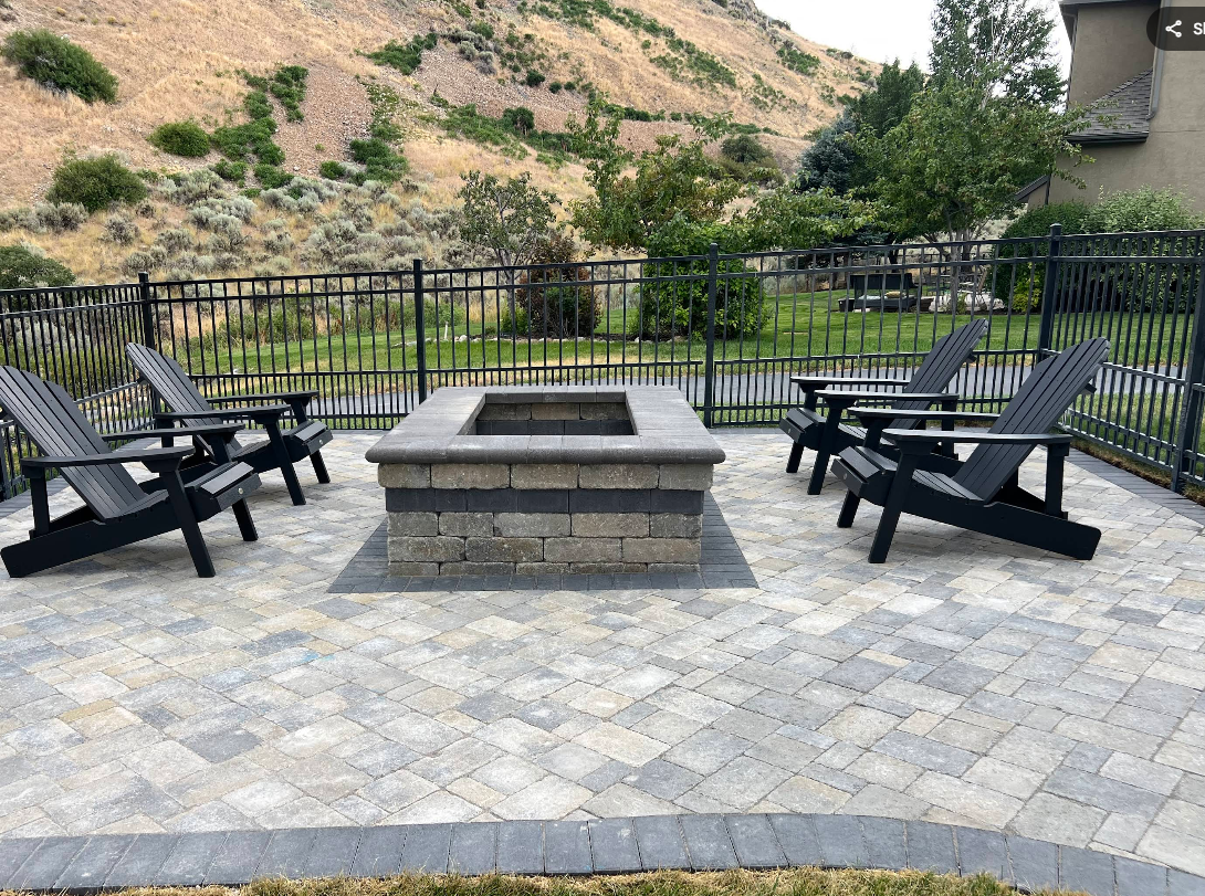 Patio with a fire pit surrounded by black Adirondack chairs and a black fence, with a hillside background.