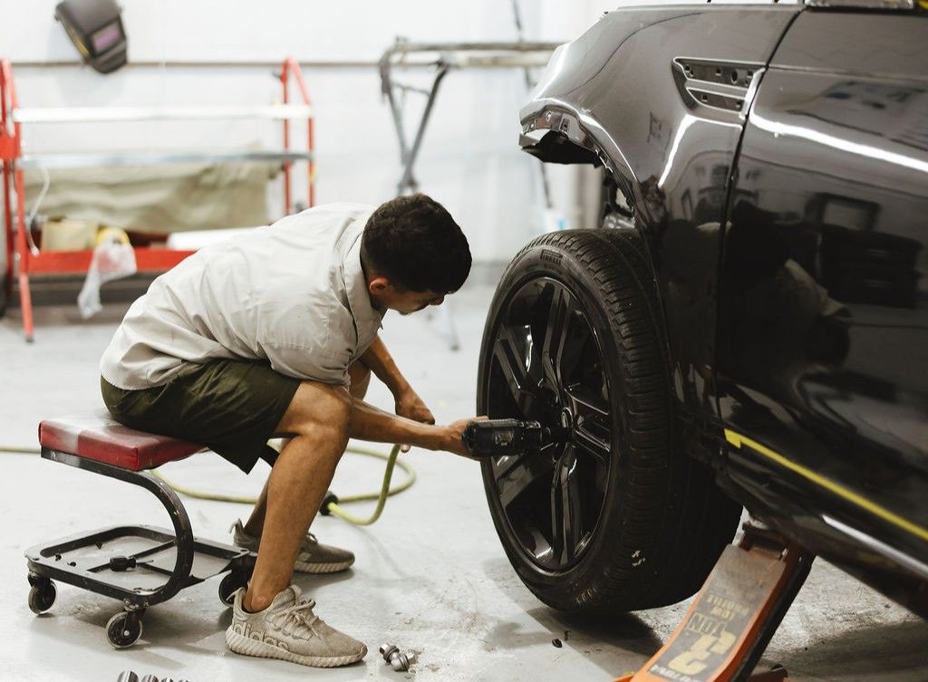 Inside an automotive workshop, a technician is using a drill to remove the lug nuts on the tire of a black car.