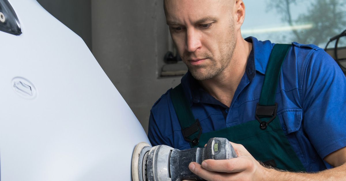 A body shop specialist using a grinder on the exterior of a car. He is wearing overalls and a blue shirt.