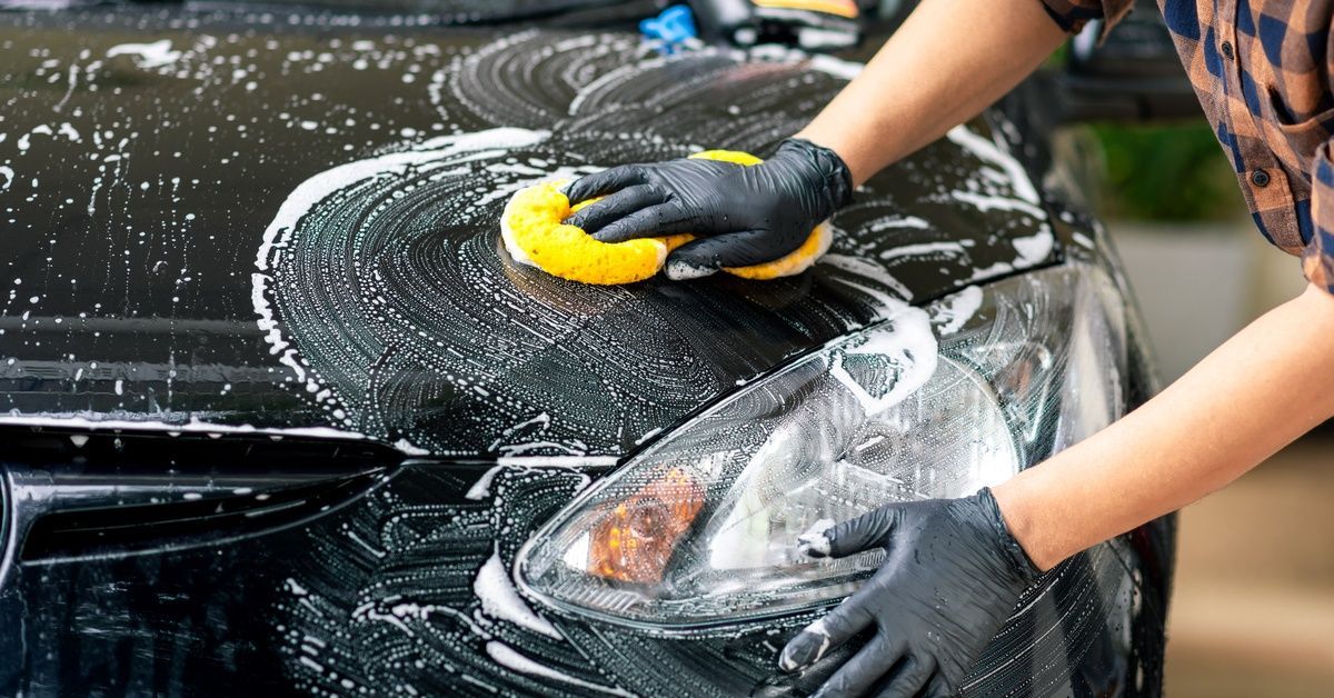A person washing the front of their car with a yellow sponge. There is white soap across the hood of