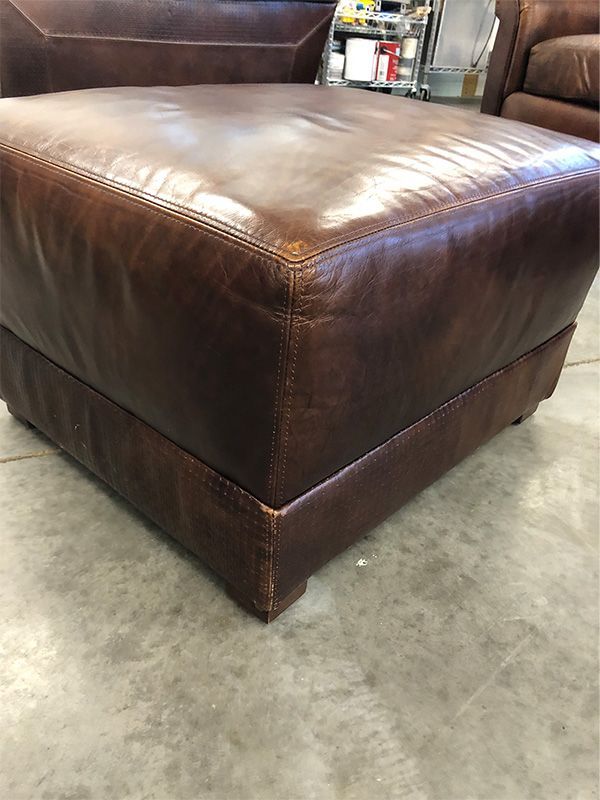 A brown leather ottoman is sitting on a concrete floor next to a chair.