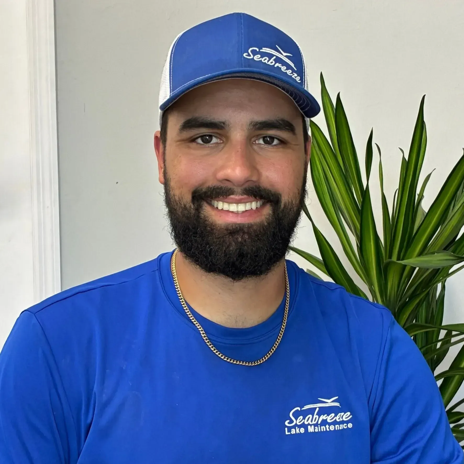 Smiling man in a blue cap and shirt, standing indoors beside a green plant.